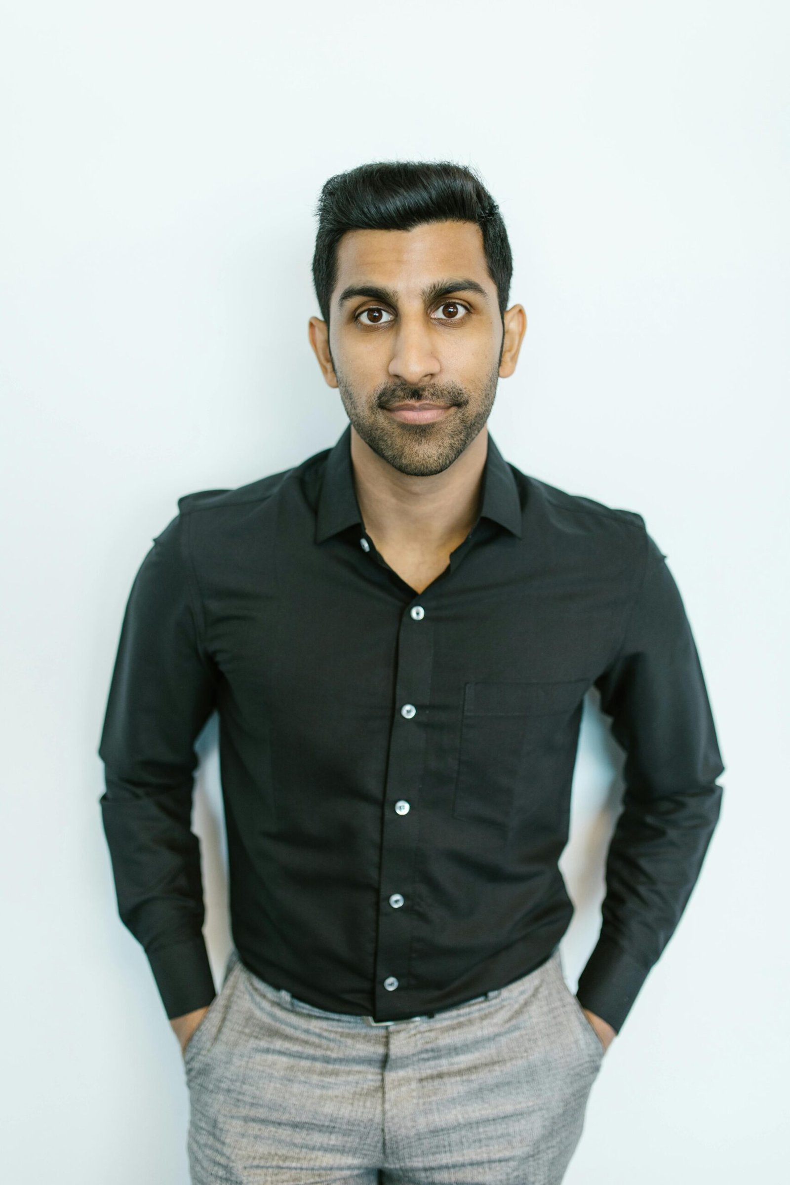 Portrait of a serious man in a black shirt with hands in pockets against a light background.